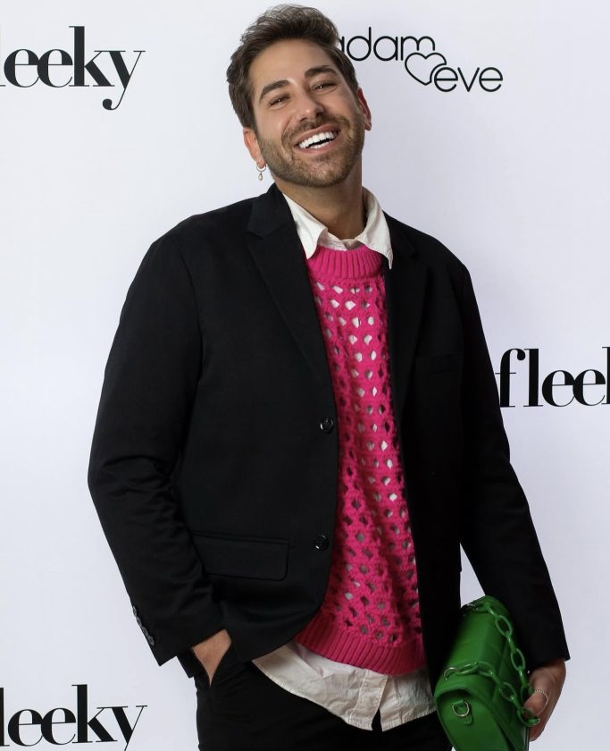 Mann (Joel) mit lächelndem Gesicht,vor einer weißen Pressewand, rosa Pullover, schwarzem Blazer und grüner Tasche.