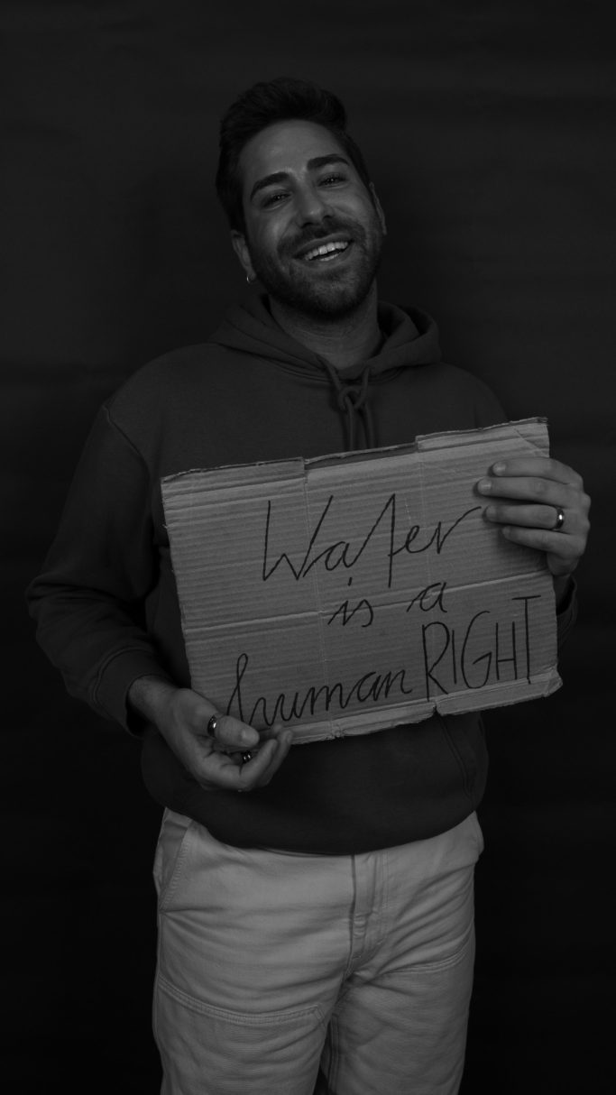 Ein lächelnder Mann (Joel) hält ein Schild mit der Aufschrift „Water is a human Right“